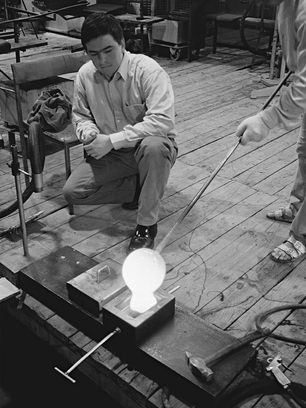 Black-and-white archive photo: a glassmaker blows a glowing bulb shape into the mould of the Block Lamp.