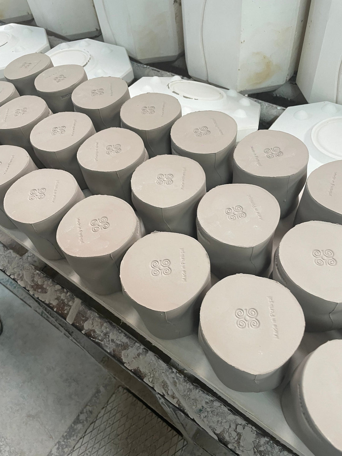 Freshly cast, unfired candle vessels of recycled clay marked Harriet Allure and Made in Portugal, in front of plaster moulds.
