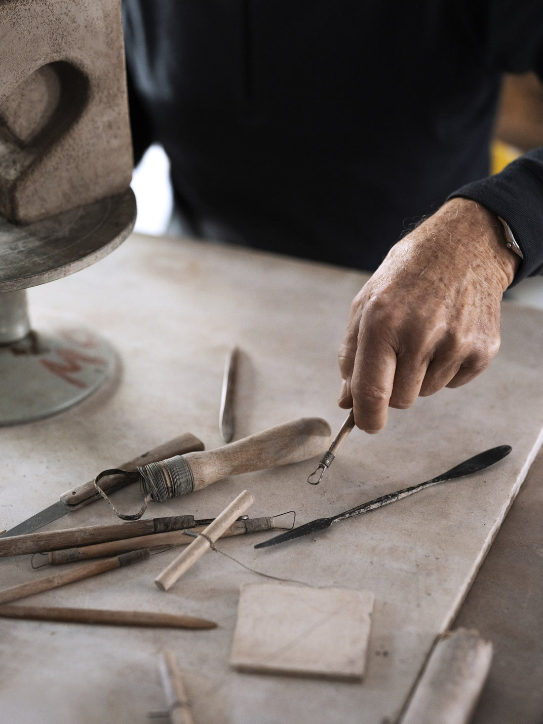 A hand picking up a modelling tool from a worktable scattered with sculpting tools.