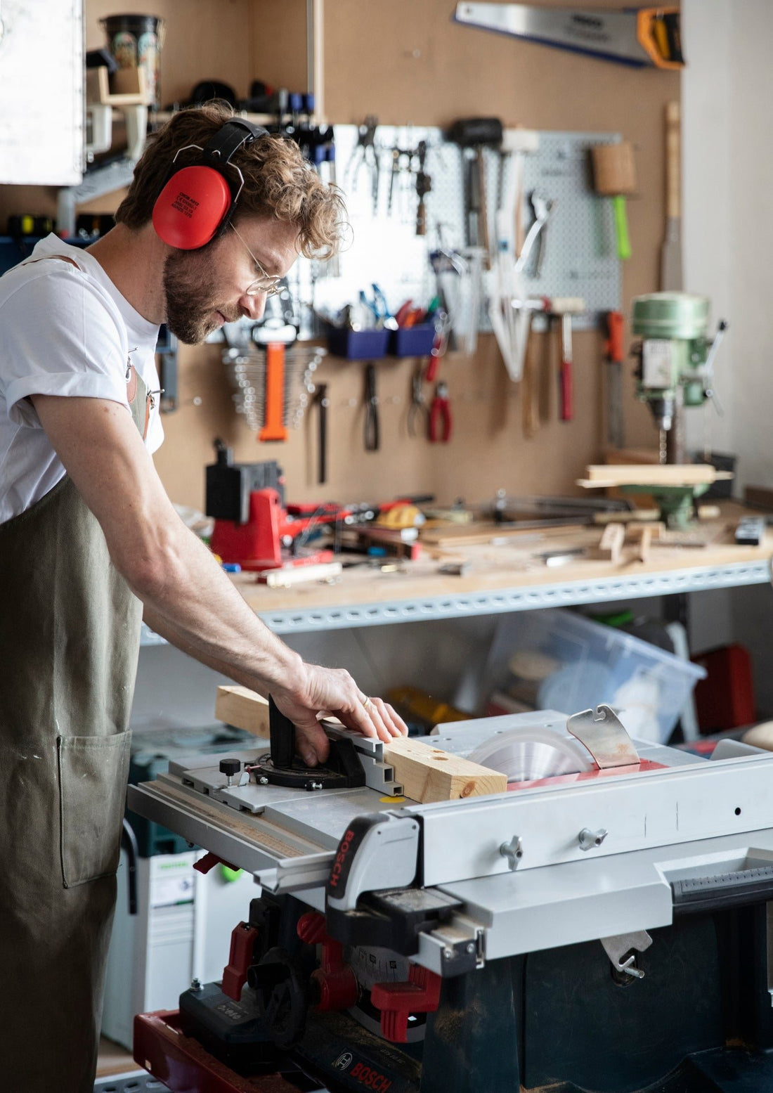 A craftsman with ear protection cuts a piece of wood on the table saw, workbench and tool wall behind.