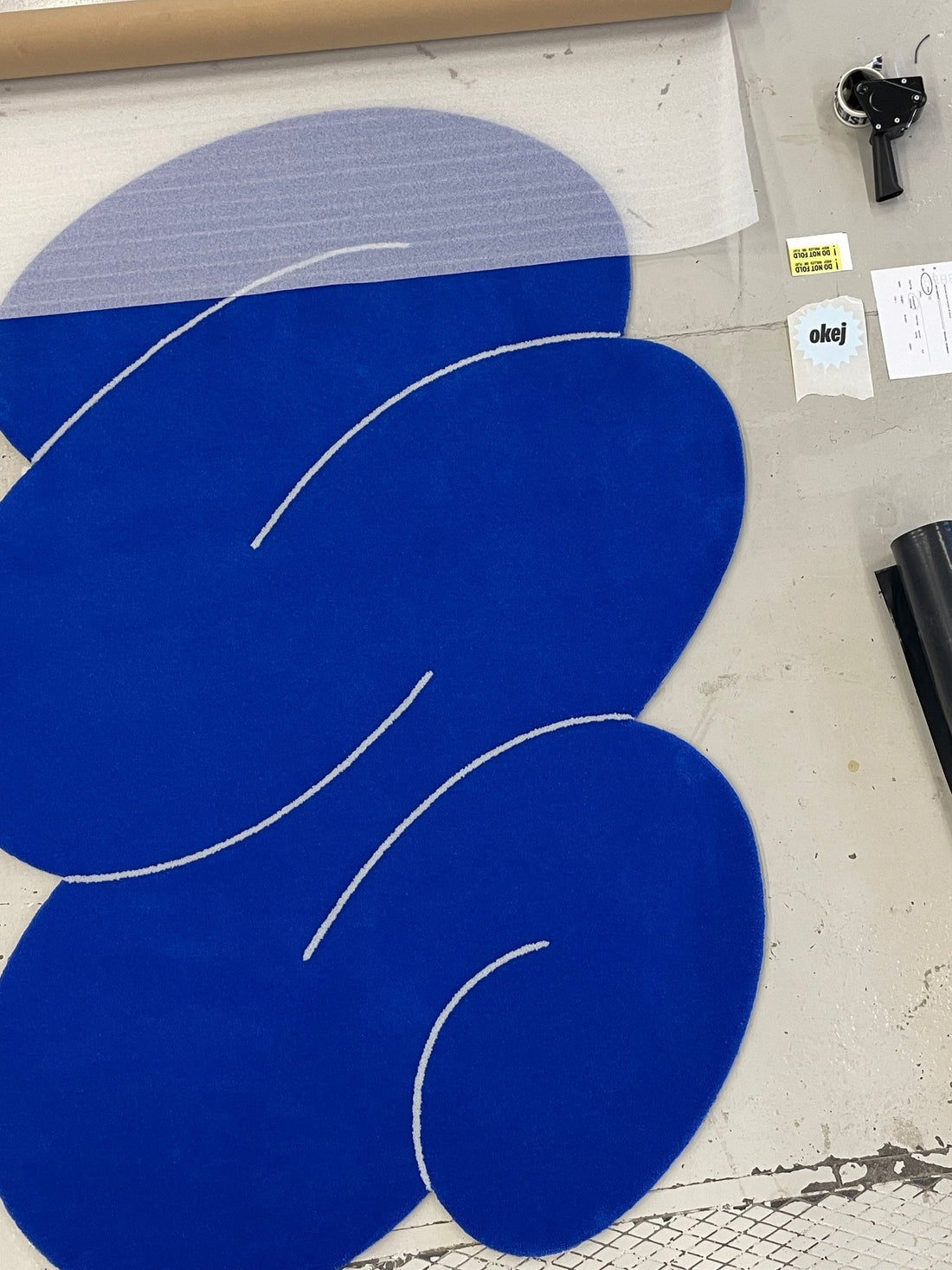 A finished tufted Neptune Squiggle rug on the workshop floor, half wrapped in protective film, Okej labels beside it.