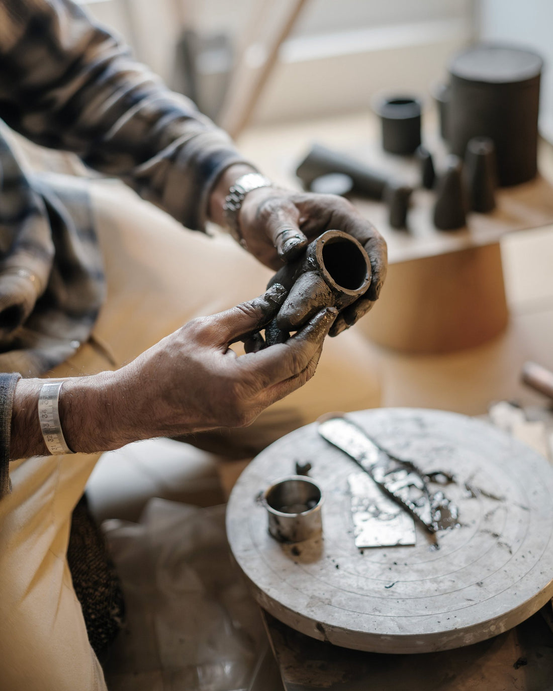 Clay-covered hands shape a small ceramic piece on the wheel, tools and finished dark objects beside it.
