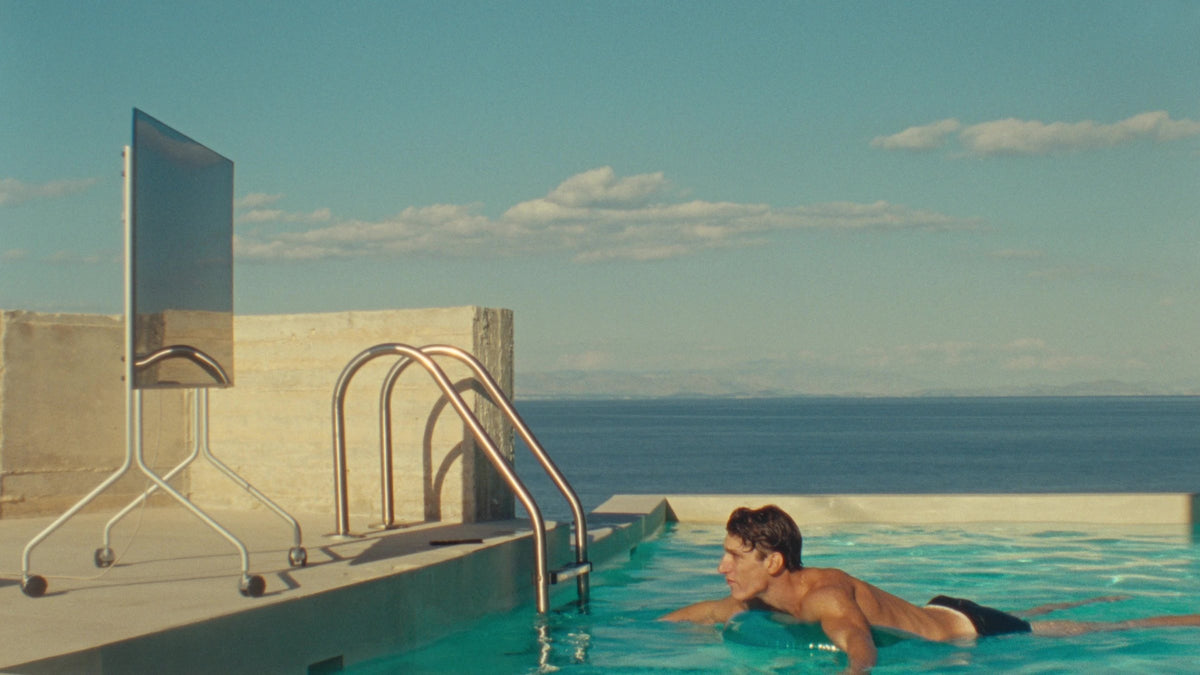 A Pedestal Moon TV stand on a concrete terrace by the sea, a man swimming in the pool beside it, still from the film Objects of Desire